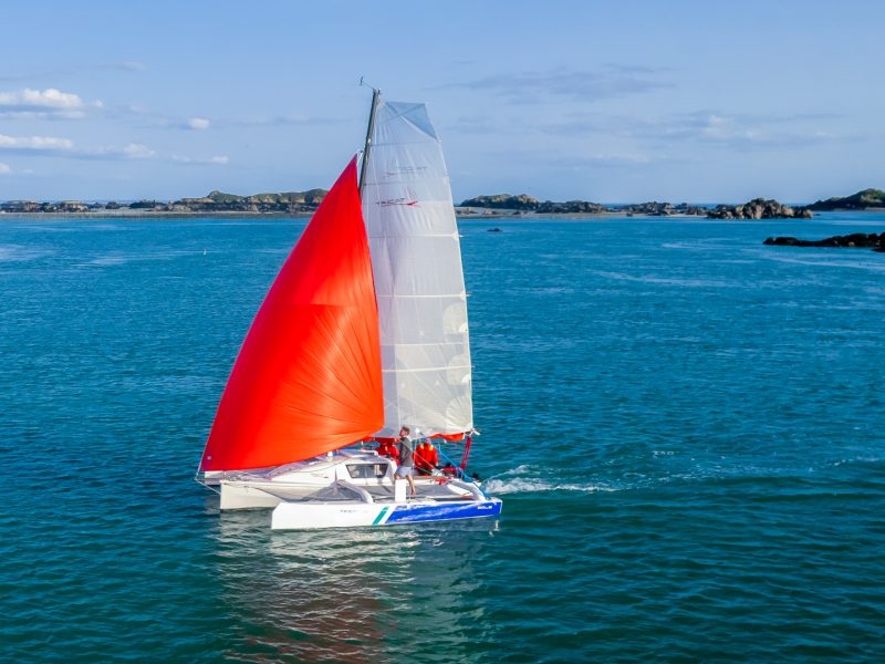 Tricat 6.90 with red foresail sailing across a bay in light winds